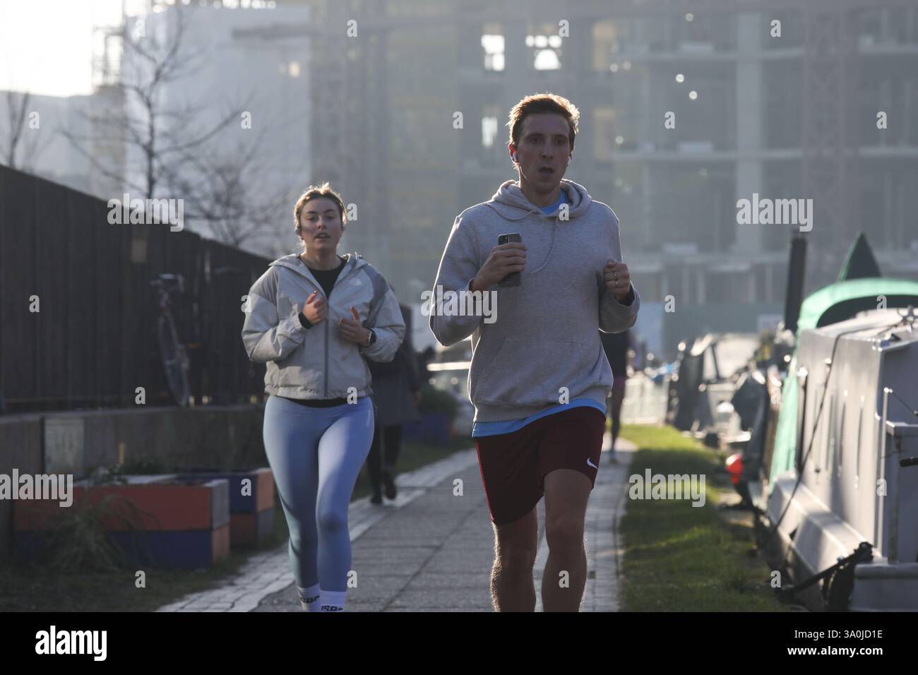 Un corridore si allena lungo il sentiero del Regent's Canal al sole mattutino di primavera. Le barche della casa costeggiano il canale con i lavori di costruzione alle spalle. Foto Stock