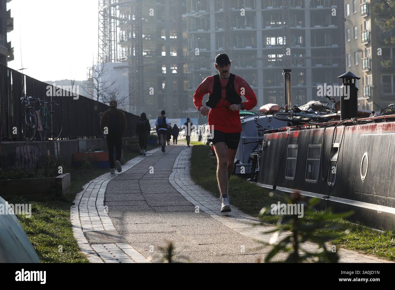 Un corridore si allena lungo il sentiero del Regent's Canal al sole mattutino di primavera. Le barche della casa costeggiano il canale con i lavori di costruzione alle spalle. Foto Stock