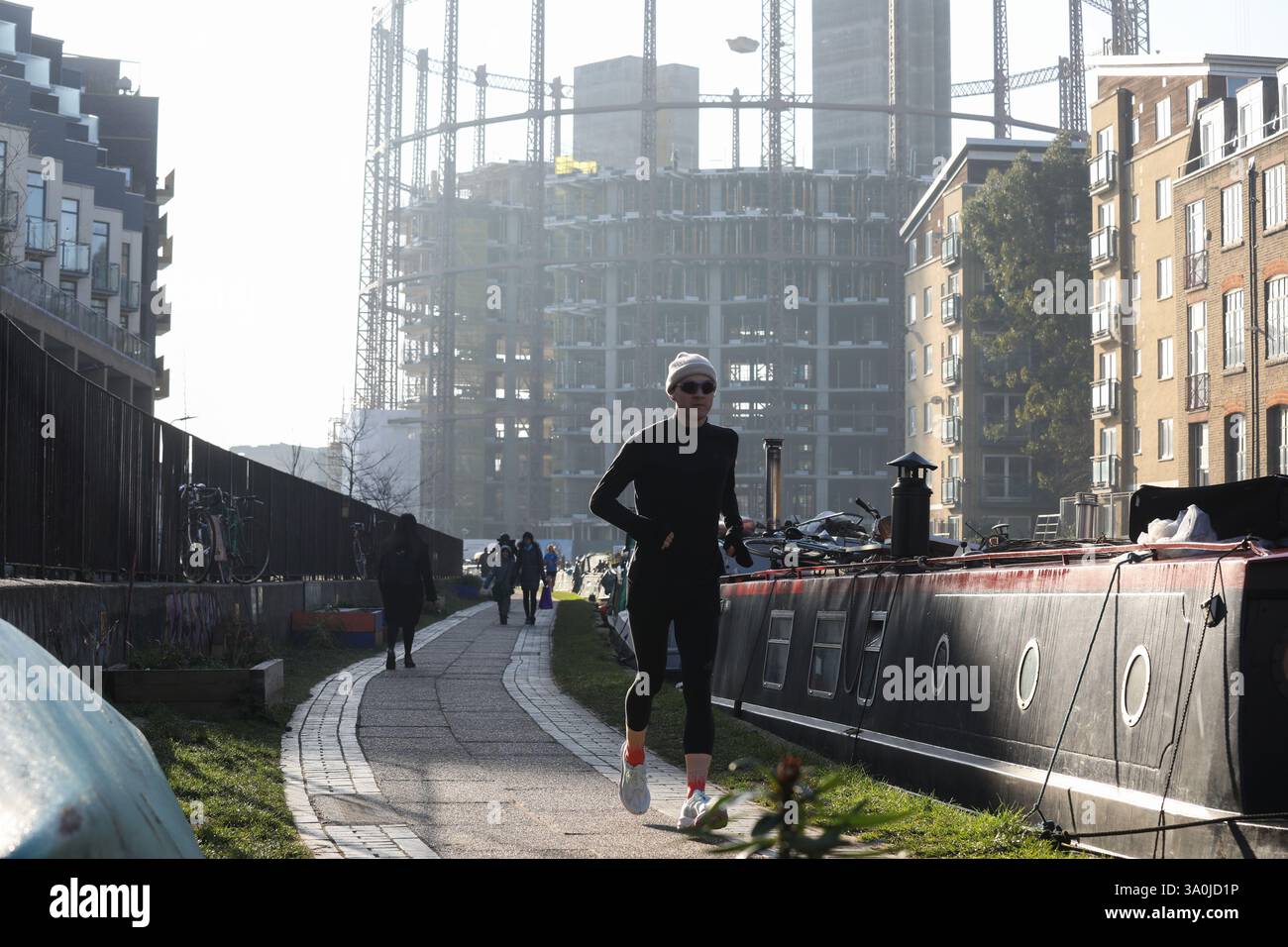 Un corridore si allena lungo il sentiero del Regent's Canal al sole mattutino di primavera. Le barche della casa costeggiano il canale con i lavori di costruzione alle spalle. Foto Stock