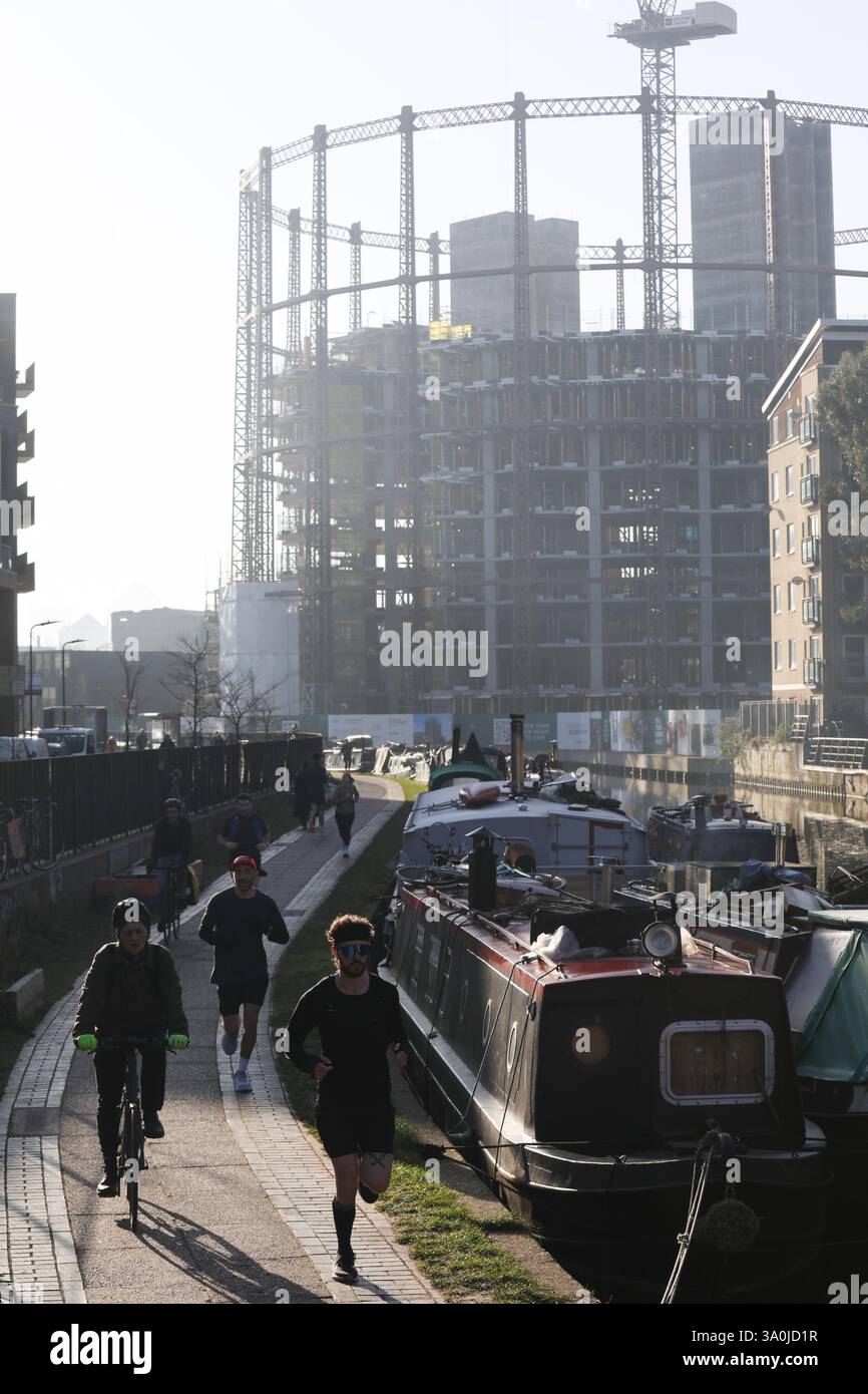 Un corridore si allena lungo il sentiero del Regent's Canal al sole mattutino di primavera. Le barche della casa costeggiano il canale con i lavori di costruzione alle spalle. Foto Stock