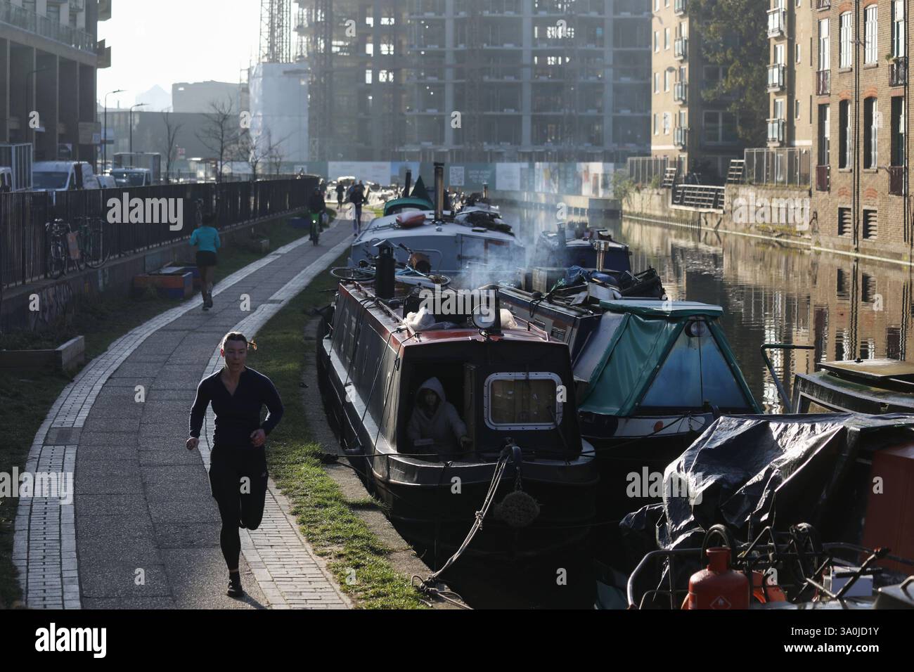Un corridore si allena lungo il sentiero del Regent's Canal al sole mattutino di primavera. Le barche della casa costeggiano il canale con i lavori di costruzione alle spalle. Foto Stock