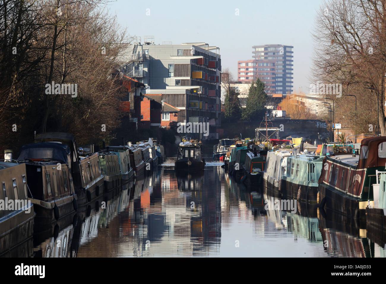 Una casa barca si fa strada lungo il Regent's Canal al sole del mattino presto. Il canale è fiancheggiato da barche che si riflettono nell'acqua. Foto Stock