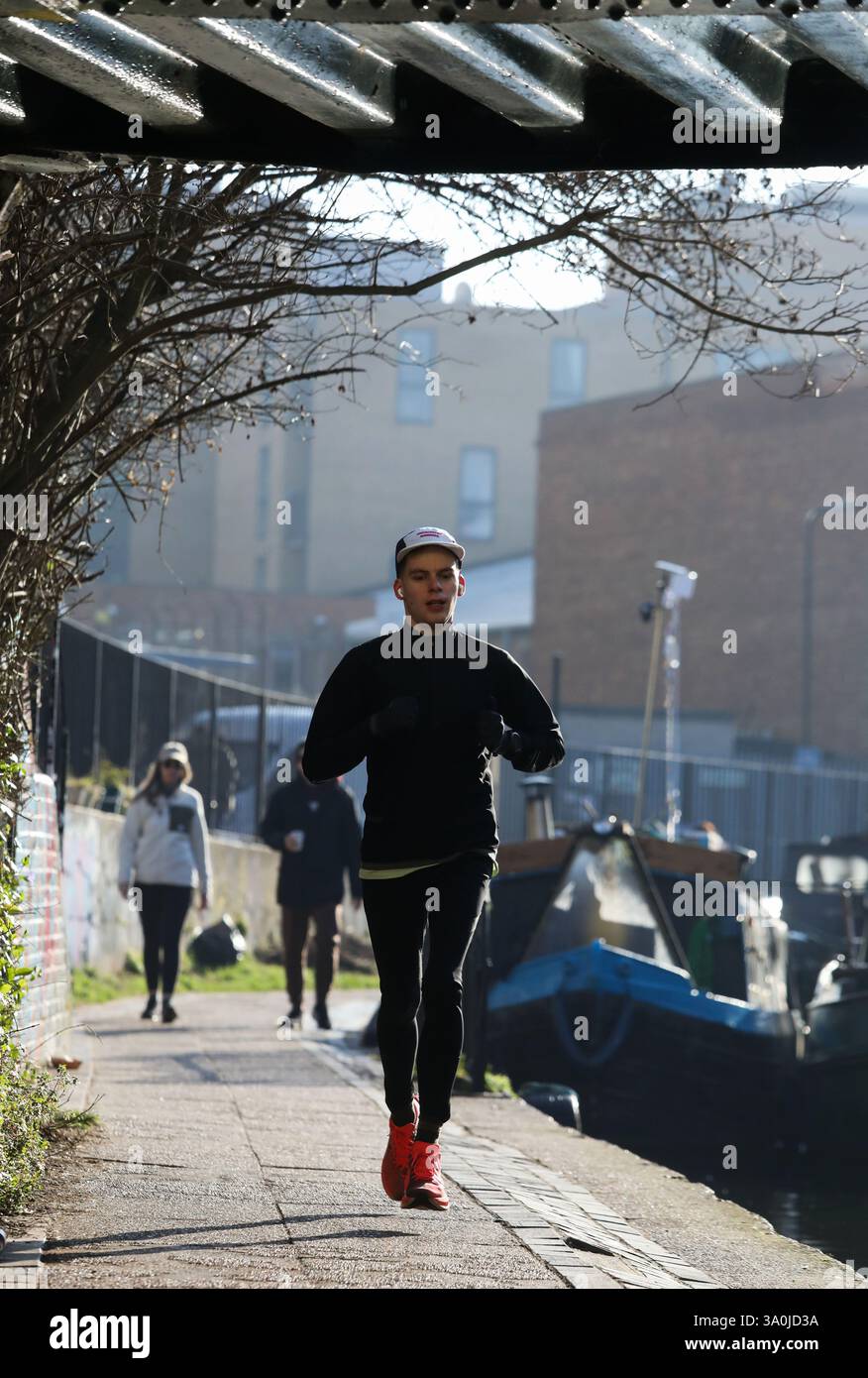 Un corridore si allena lungo il sentiero del Regent's Canal al sole mattutino di primavera. Le barche della casa costeggiano il canale e il sole si riflette nel ponte. Foto Stock