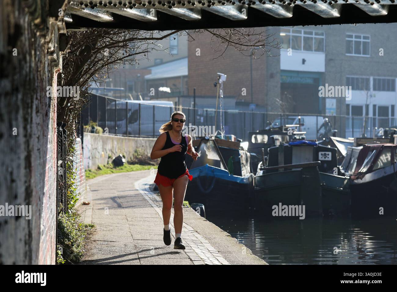 Un corridore si allena lungo il sentiero del Regent's Canal al sole mattutino di primavera. Le barche della casa costeggiano il canale e il sole si riflette nel ponte. Foto Stock