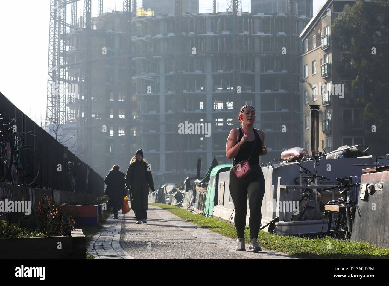 Un corridore si allena lungo il sentiero del Regent's Canal al sole mattutino di primavera. Le barche della casa costeggiano il canale con i lavori di costruzione alle spalle. Foto Stock