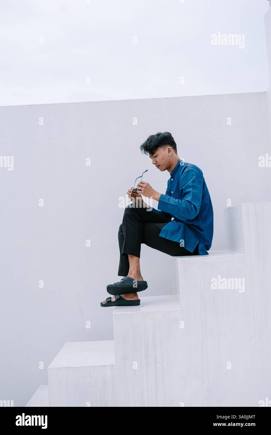 Un giovane premuroso siede su una scala bianca minimalista, con gli occhiali da vista. È vestito con un abbigliamento casual ma elegante, che incarna uno stile moderno Foto Stock