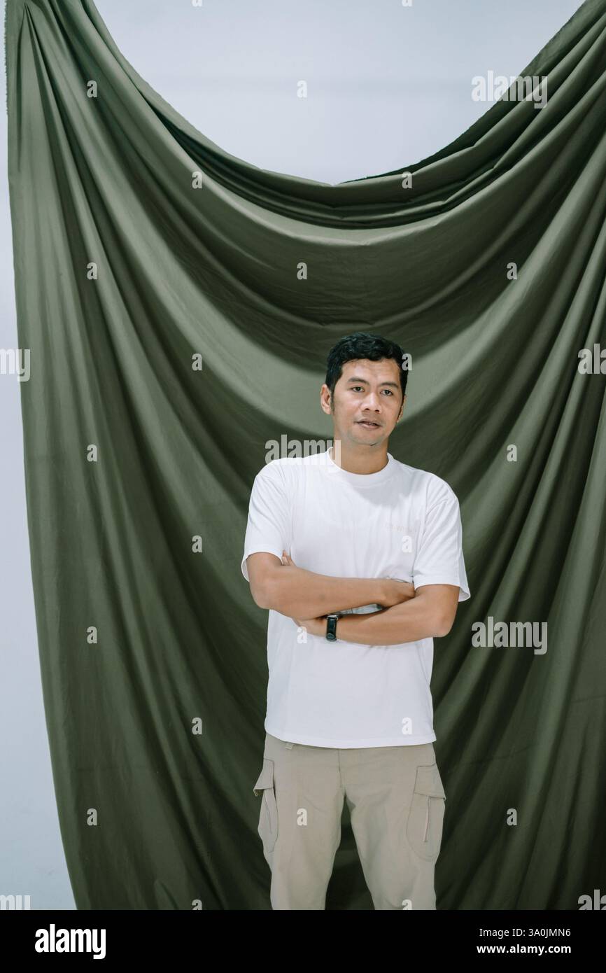 Ritratto in studio di un uomo asiatico in piedi con fiducia con le braccia incrociate su uno sfondo drappeggiato. L'uomo è vestito casualmente e posa con un neut Foto Stock