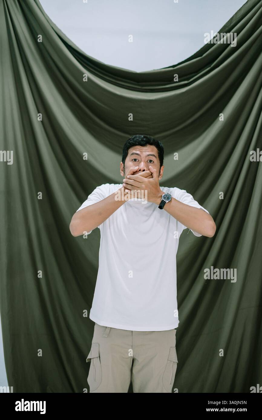 Un uomo con un'espressione sorpresa si trova di fronte a uno sfondo verde scuro. Sta coprendo la bocca con le mani, suggerendo il concetto di ke Foto Stock