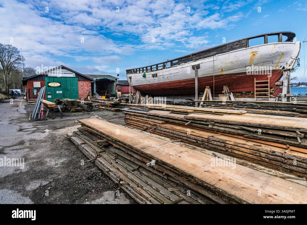 Tavole di legno e una grande nave in restauro in un caratteristico cantiere navale in riva al mare. L'area espone attrezzi e capannoni, che riflettono gli artigiani tradizionali Foto Stock