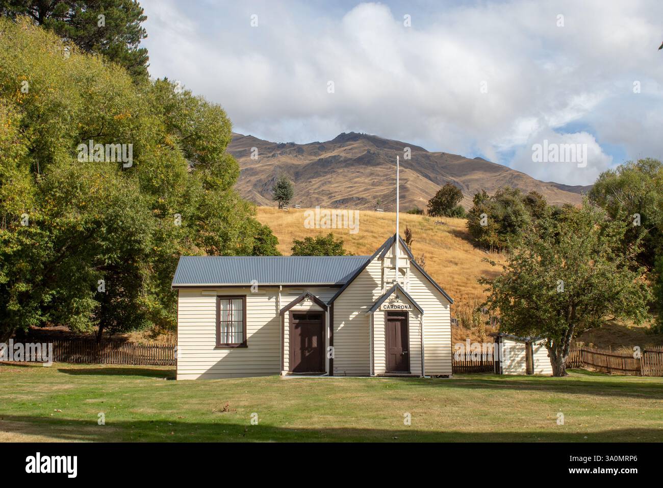 La chiesa si erge tranquillamente in un campo erboso, le montagne si innalzano sullo sfondo. Edificio bianco con porte scure, un palo alto. Foto Stock