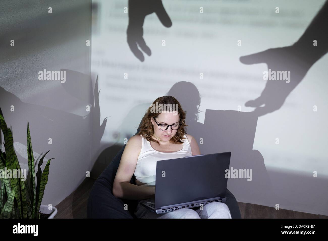 Una donna si è concentrata intensamente sul suo notebook mentre le inquietanti ombre incombono su di lei, descrivendo i gravi rischi delle minacce online Foto Stock