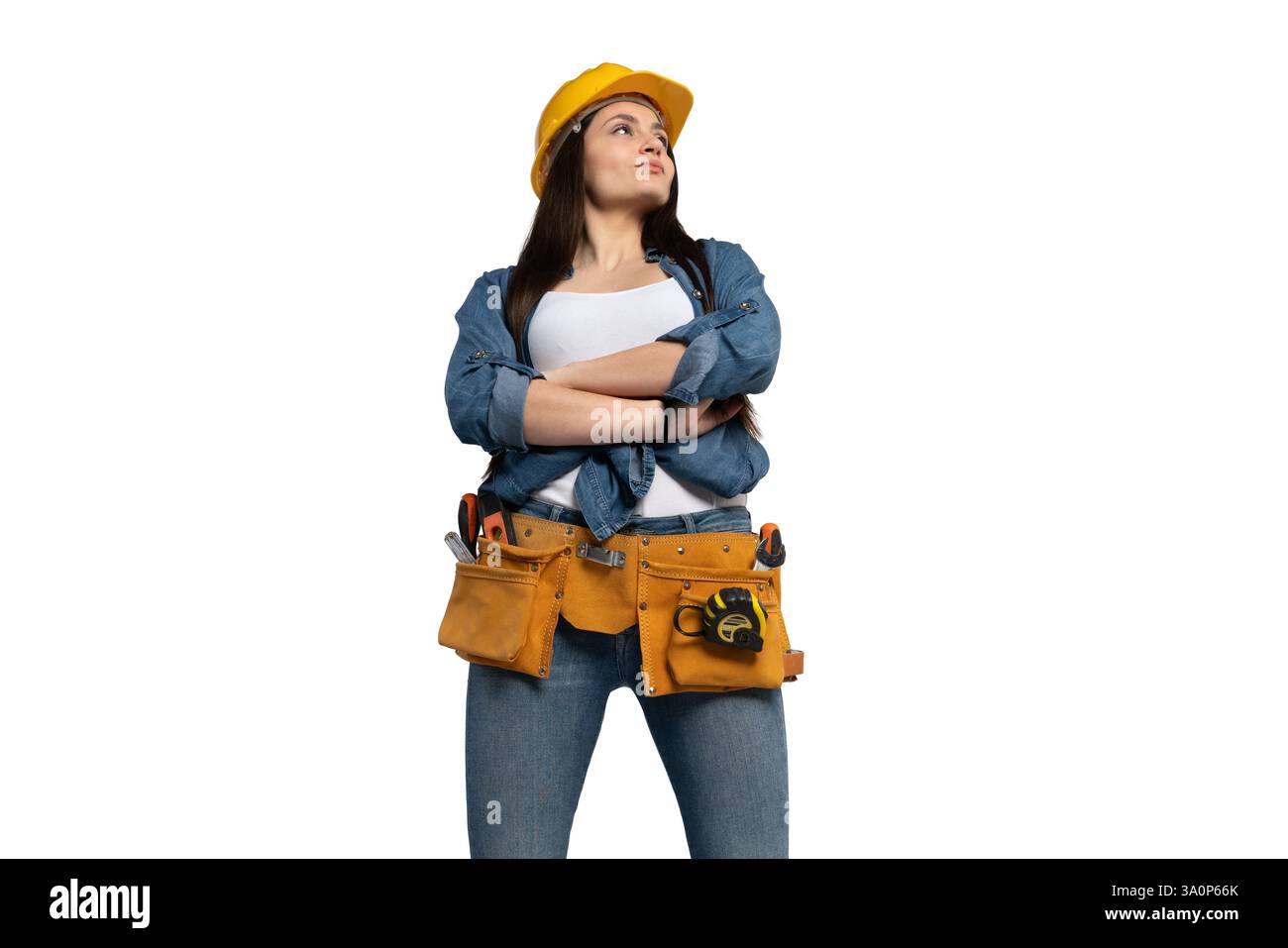 Una lavoratrice edile forte e sicura di sé, che indossa un elmetto giallo e una cintura portautensili, sta con le braccia incrociate, guardando verso l'alto con determinazione Foto Stock