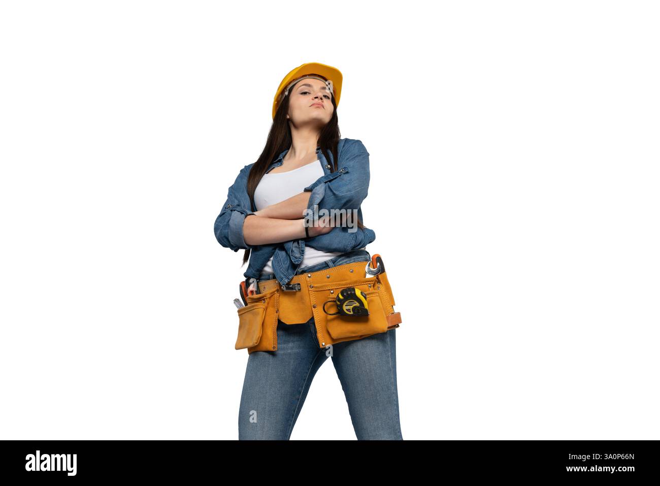 Una lavoratrice edile sicura di sé con un elmetto giallo e una cintura portautensili si staglia con le braccia incrociate, guardando verso il basso con un'espressione audace. Questo imag Foto Stock