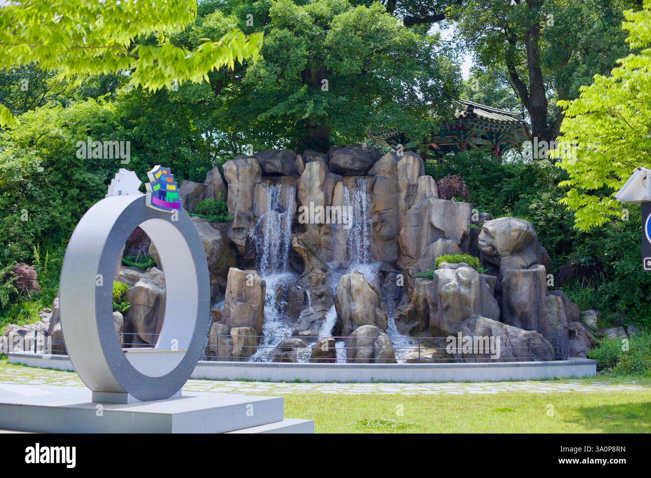 Iksan, Corea del Sud - 28 maggio 2021: Cascata artificiale circondata da vegetazione lussureggiante e architettura coreana tradizionale in riva al fiume Foto Stock