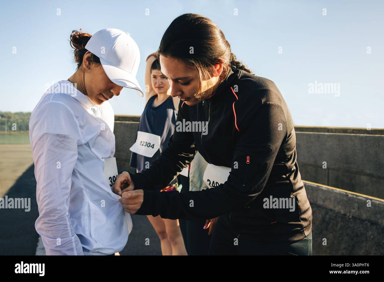 Tre amici si stanno preparando per una maratona, con uno che aiuta l'altro ad attaccare un numero di gara alla sua maglietta. Si trovano su una pista soleggiata all'aperto, mostra Foto Stock