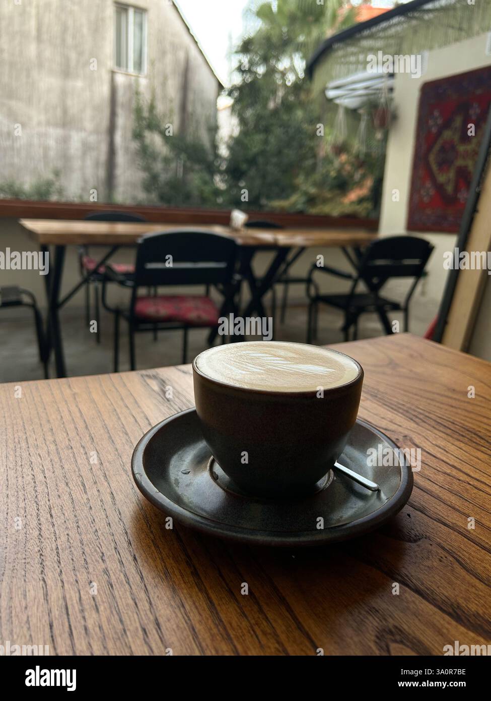 Una tazza di caffè in ceramica con latte art siede su un tavolo di legno in un caffè all'aperto con tavoli e sedie vuote sullo sfondo. L'impostazione include Foto Stock