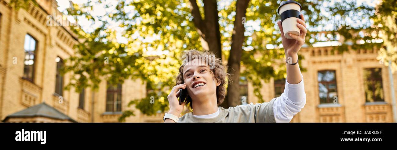 Lo studente elegante parla con gioia al telefono con un drink in mano in un ambiente vivace del campus. Foto Stock
