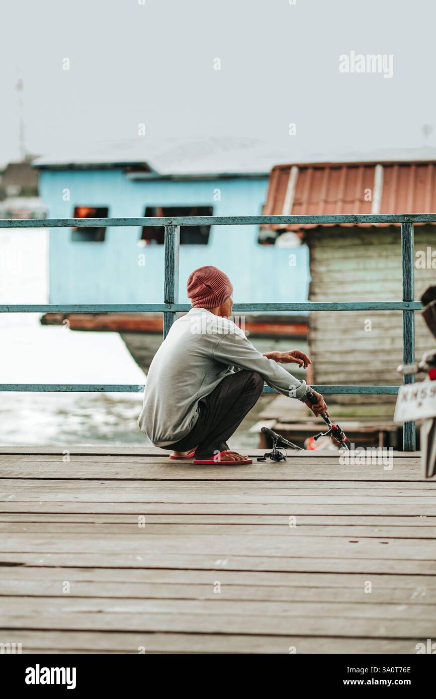 Un pescatore siede su un molo di legno, godendosi il fiume calmo in un villaggio galleggiante. Una vita semplice vicino all'acqua, piena di storie. Foto Stock