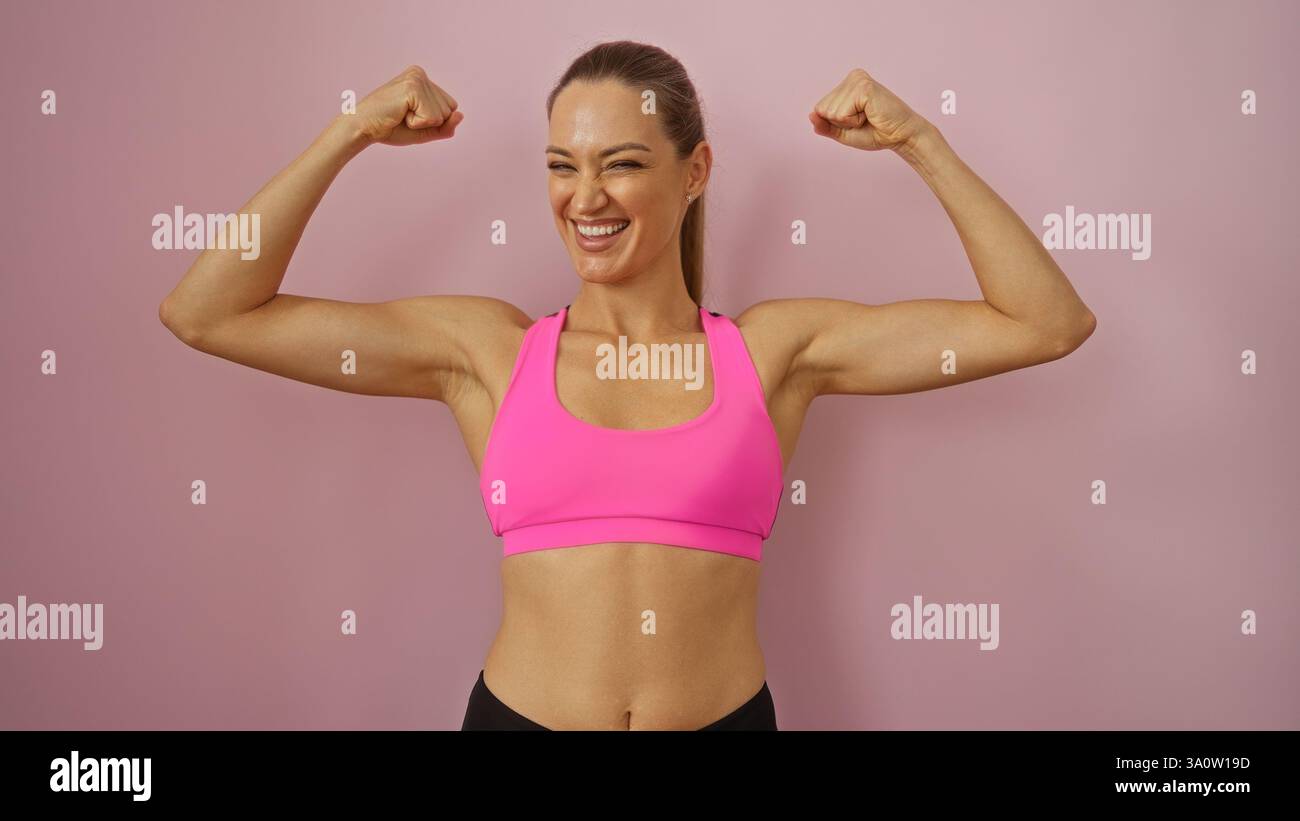 Donna che si flette con sicurezza su sfondo rosa, mostrando forza e potenza in un vivace abbigliamento sportivo, trasmettendo salute e positività. Foto Stock