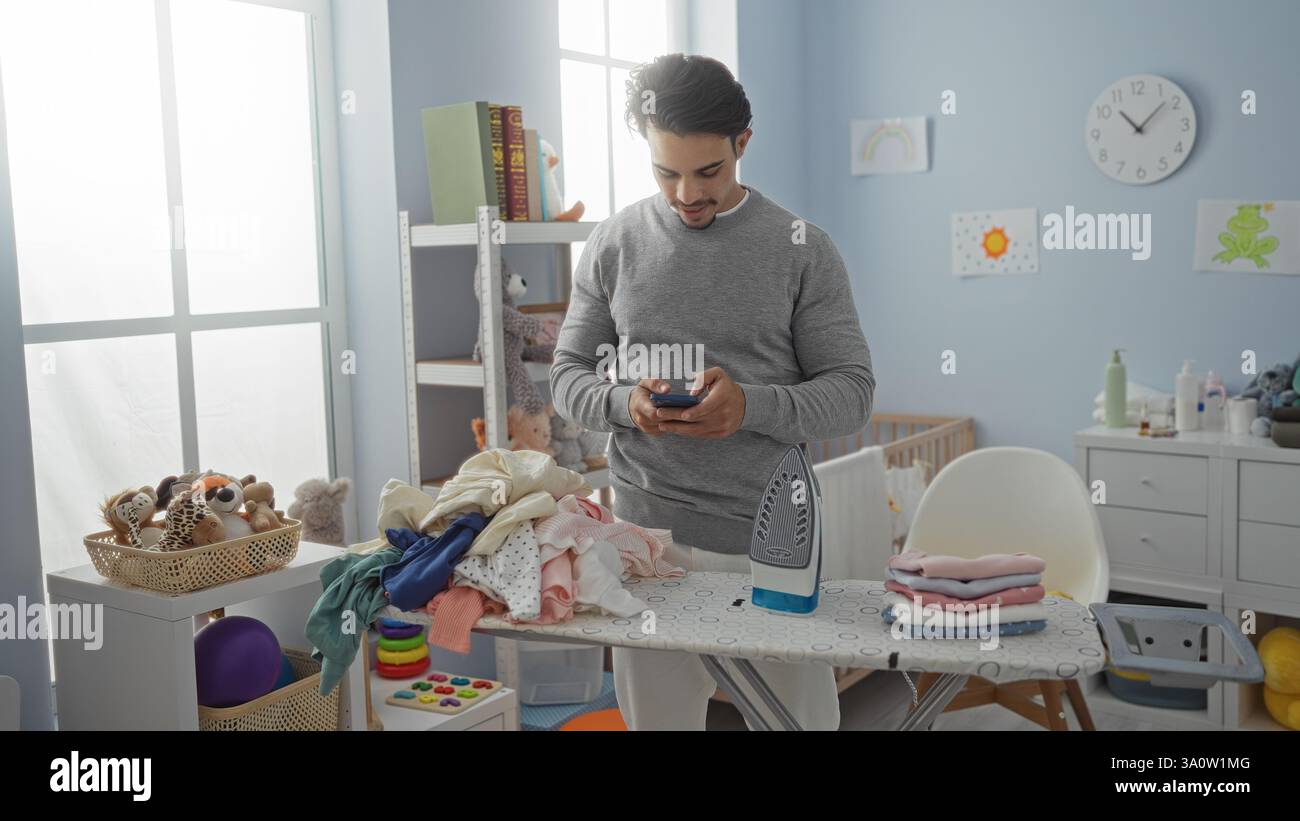 Giovane ispanico in una lavanderia a casa che controlla il telefono mentre stirava i vestiti, circondato da giocattoli per bambini e culla, trasportando un multitasking Foto Stock