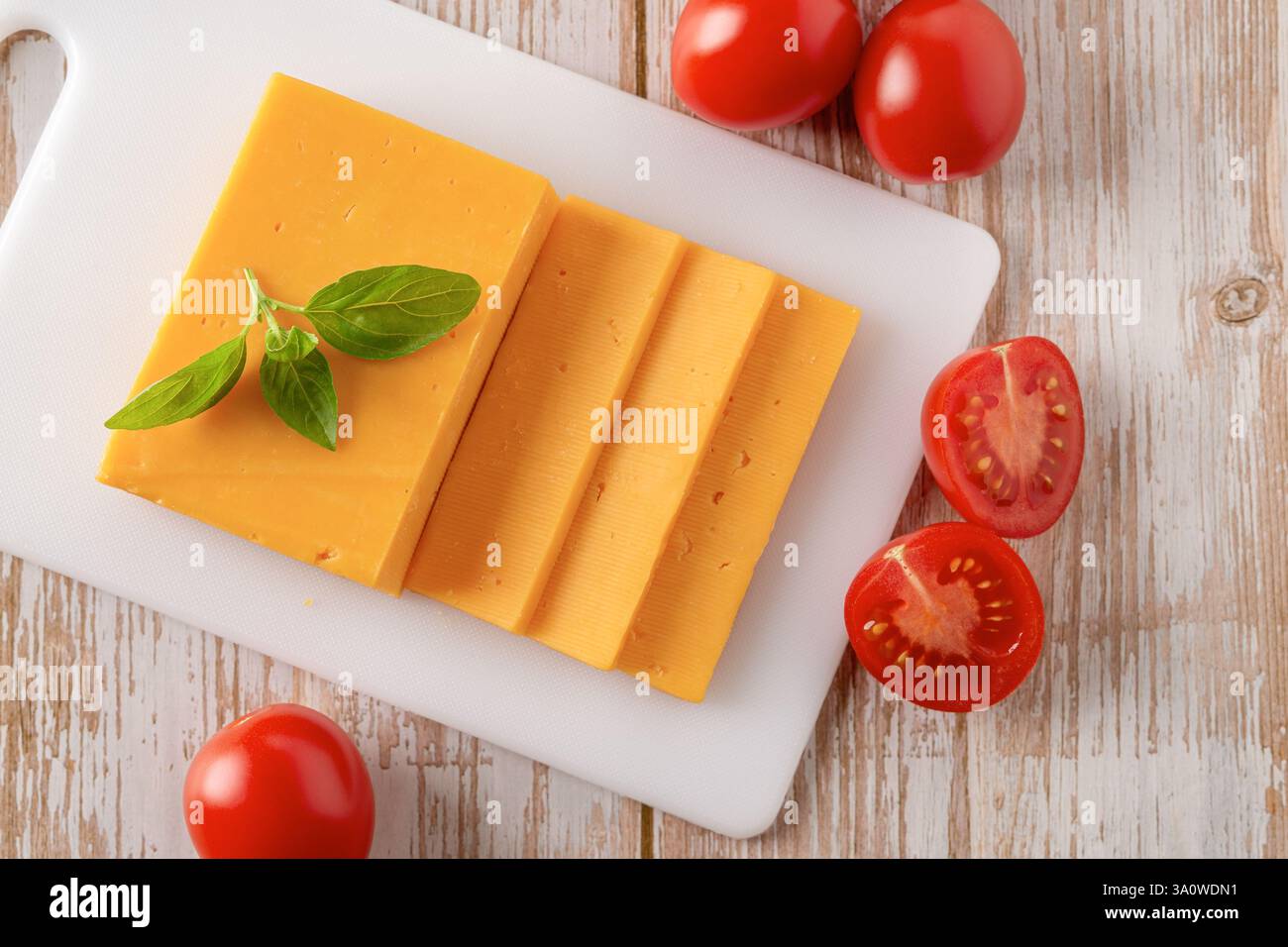 Fette di formaggio Sheddar e primi piani di pomodori. Formaggio d'arancia intero e tagliato su un tagliere bianco e pochi pomodori rossi su un tavolo in legno. Foto Stock