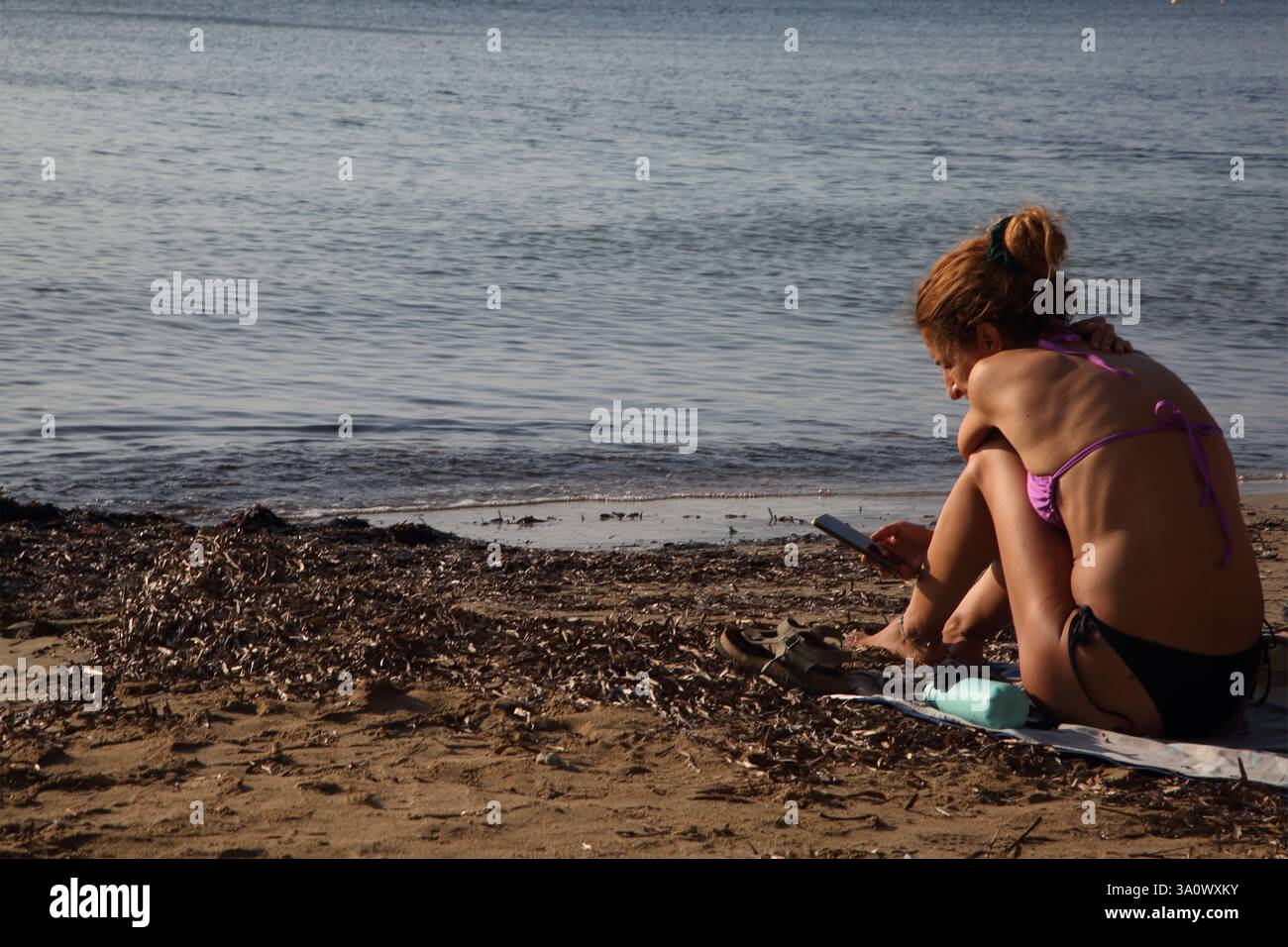 Donna che guarda un telefono cellulare seduto sulla spiaggia di Vouliagmeni, Attica Grecia Foto Stock