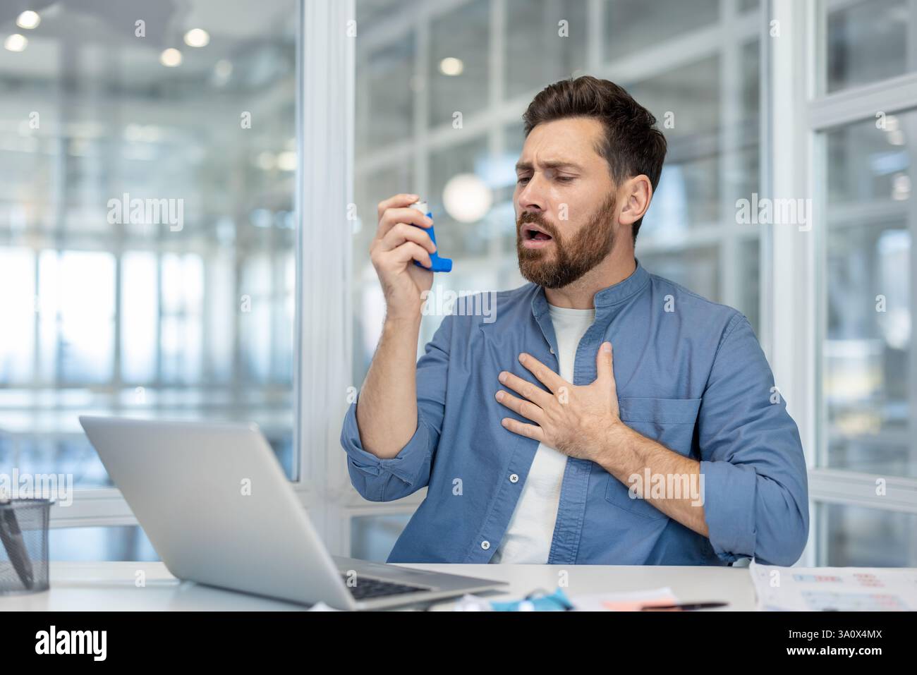 Un uomo che usa un inalatore in un ufficio, cerca di alleviare le sofferenze respiratorie mentre è seduto alla sua scrivania con un portatile nelle vicinanze. Foto Stock