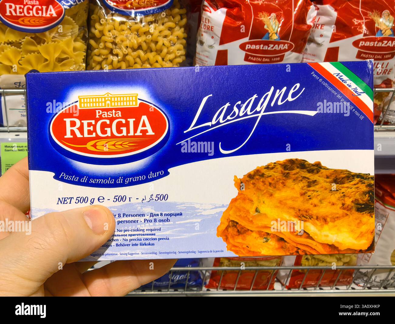 Bobruisk, Bielorussia - 22 febbraio 2025: Una mano tiene una scatola di lasagne in un vivace corridoio di alimentari pieno di vari prodotti di pasta. Foto Stock