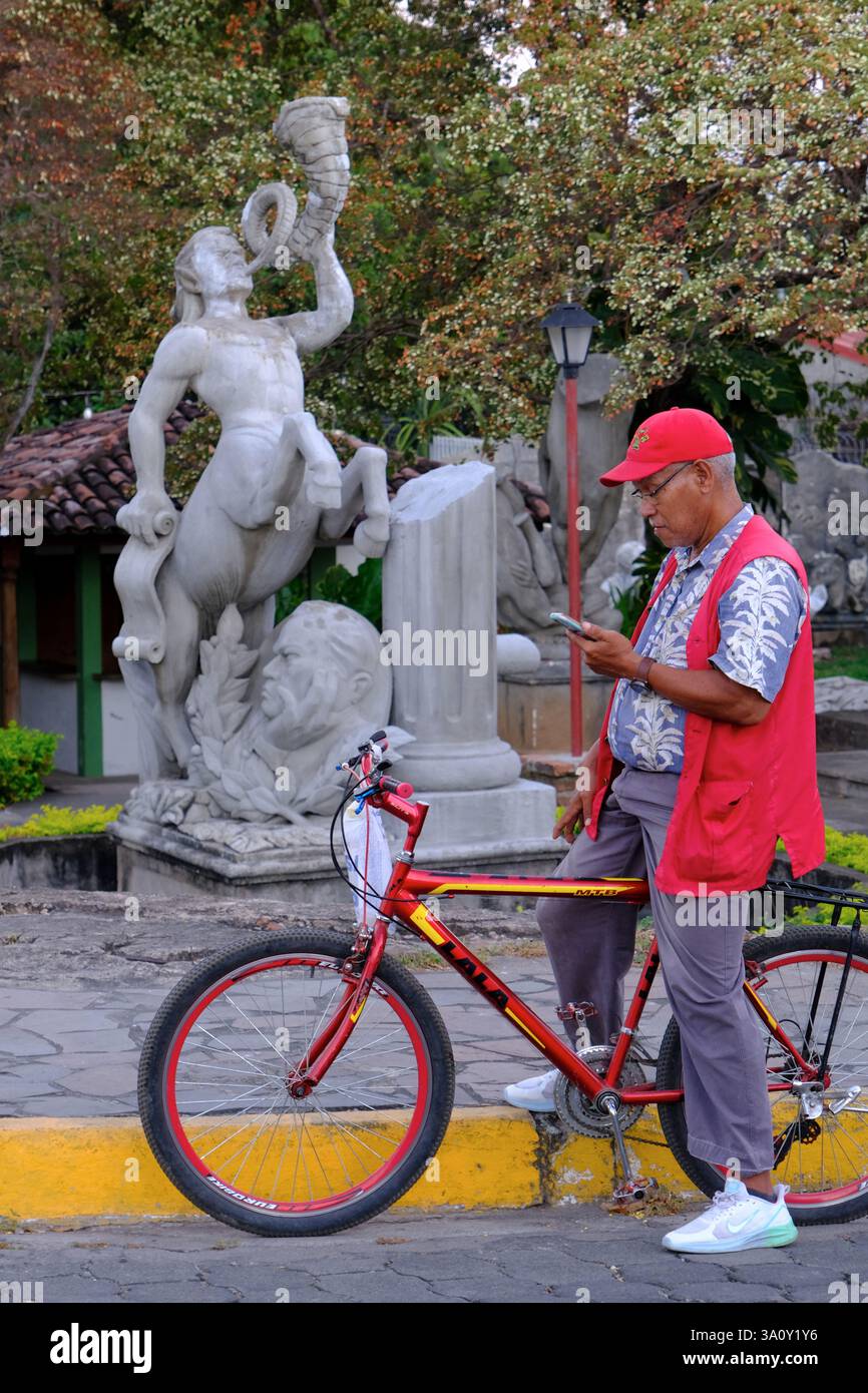 Un uomo anziano che indossa un cappello rosso e un giubbotto rosso seduto sulla sua bicicletta a controllare il suo cellulare con una statua di Centaur sul retro.Granada.Nicaragua Foto Stock
