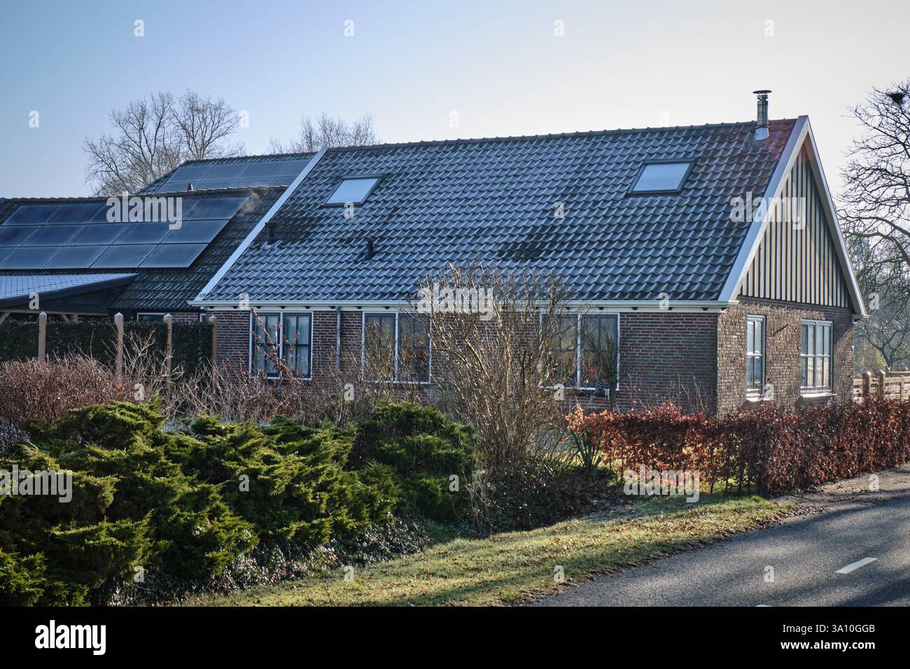 Un fienile rustico con tetto piastrellato, e dietro di esso un capannone coperto da pannelli solari, circondato da piante e alberi ricoperti di gelo sotto un cielo azzurro. Foto Stock
