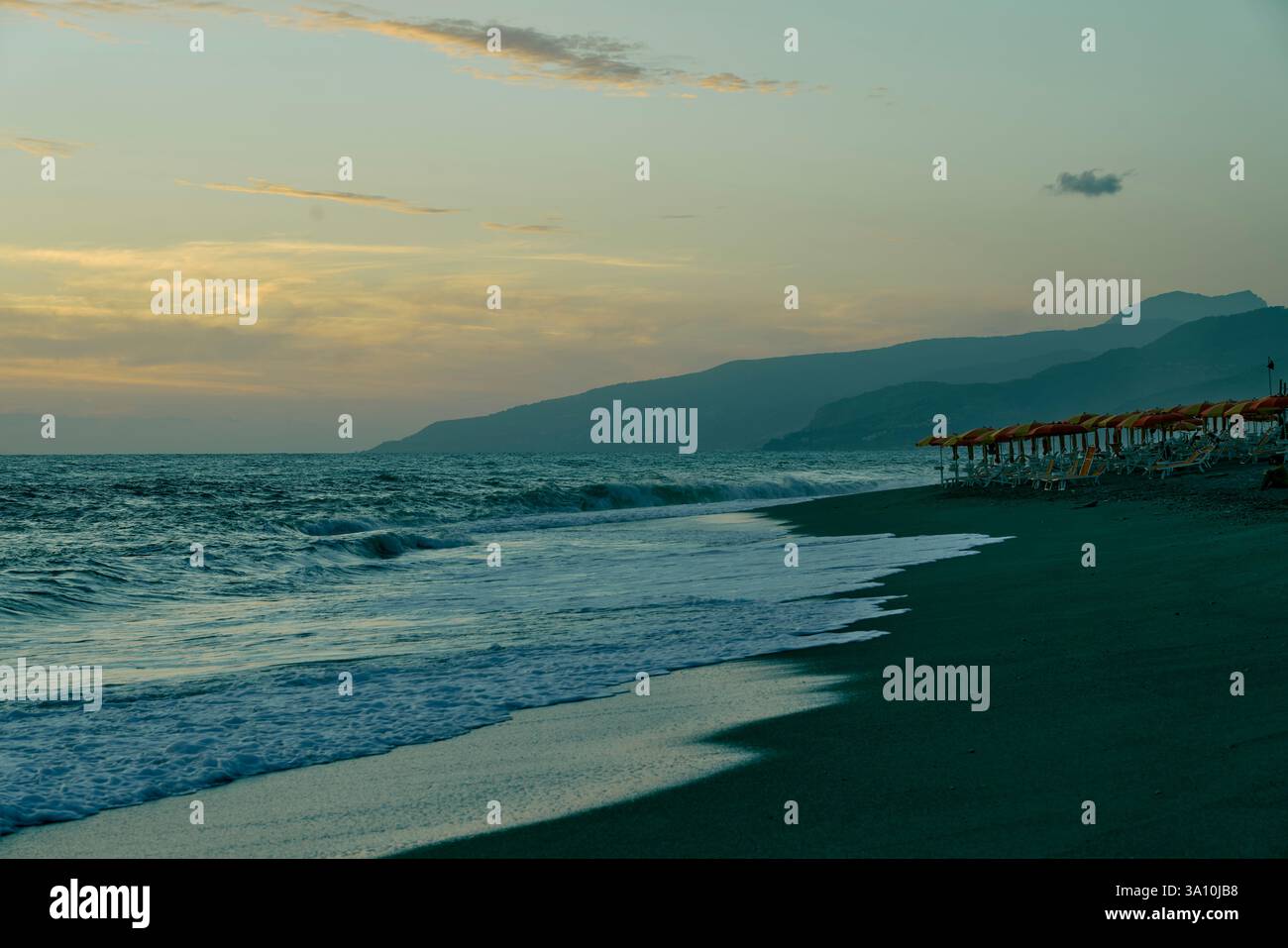 Mare con file di ombrelloni arancioni sulla riva al tramonto con montagne sullo sfondo, Paola, Calabria, Italia. Foto Stock