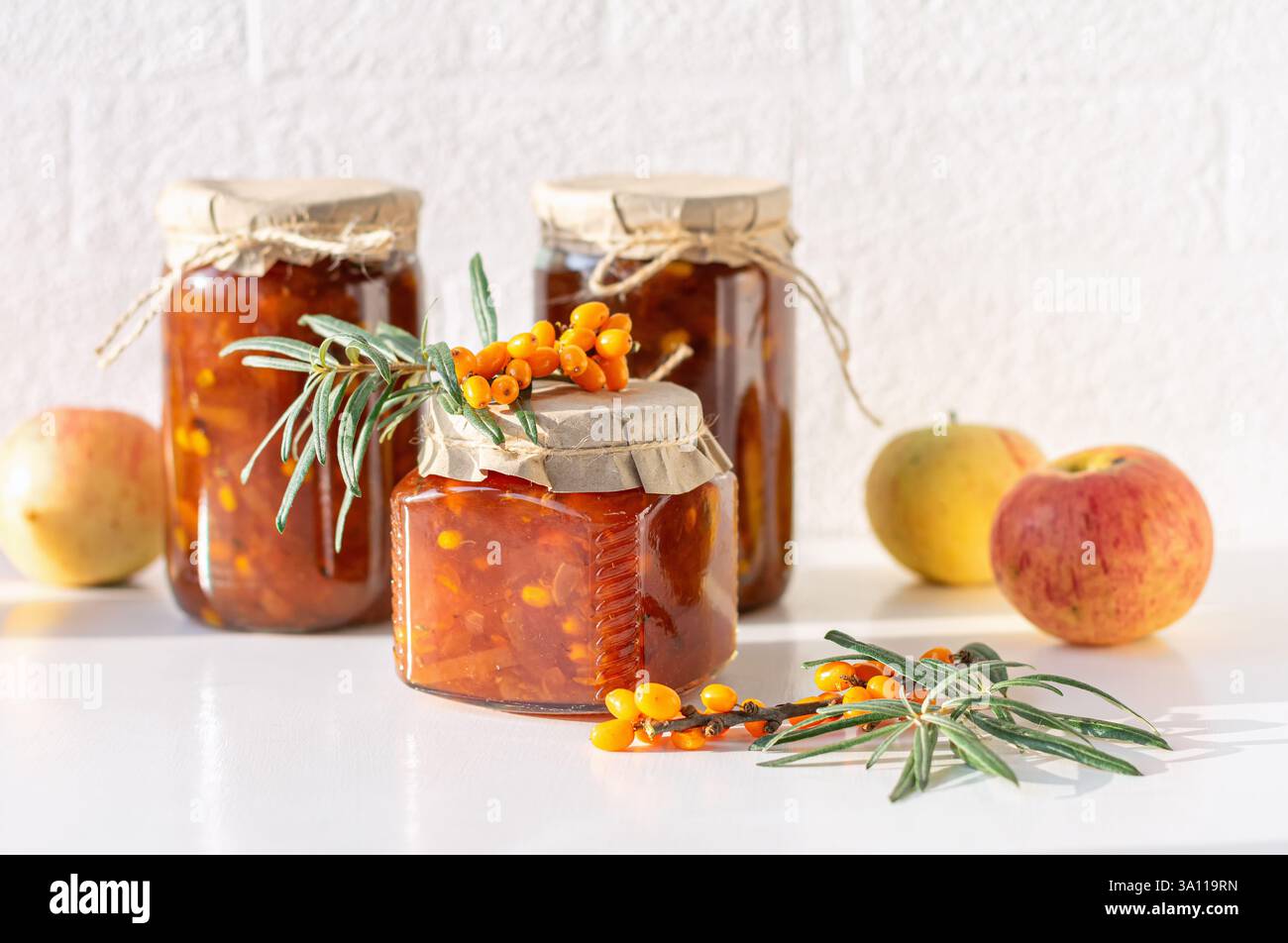 Tre vasetti di spinoso marino e marmellata di mele sono esposti su un tavolo accanto alle mele fresche e ai frutti di spinone marino. Foto Stock