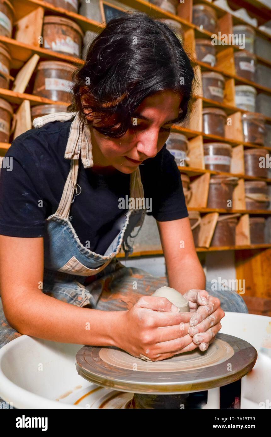 Artigiana che forma l'argilla su una ruota di ceramica, circondata da contenitori di materiali nella sua officina Foto Stock