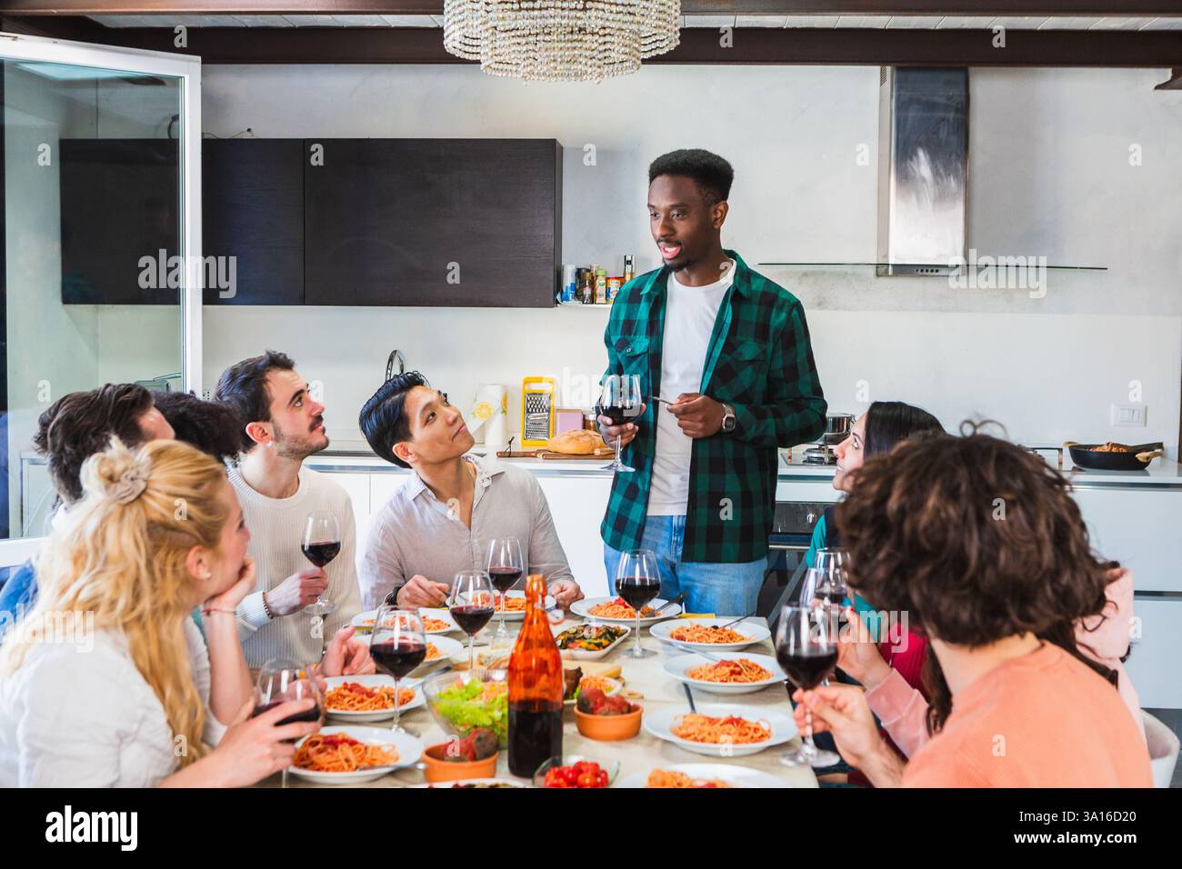 Un gruppo di amici si diverte a casa, ascoltando un giovane che fa un brindisi Foto Stock
