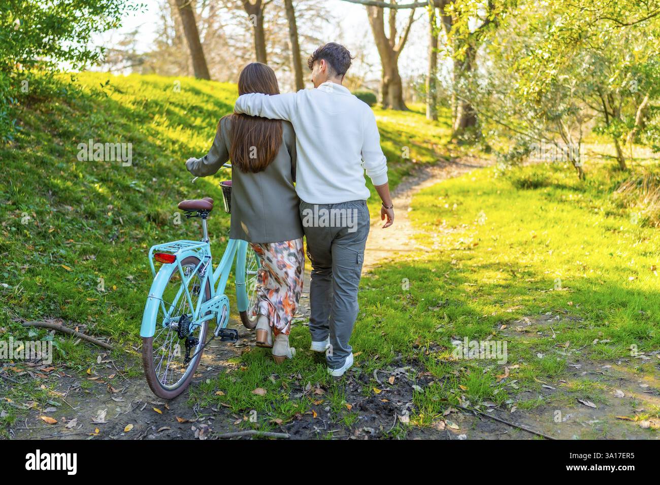 Una coppia passeggia attraverso un parco illuminato dal sole, abbracciandosi l'un l'altro mentre cammina accanto a una bicicletta su un sentiero panoramico circondato dal verde Foto Stock