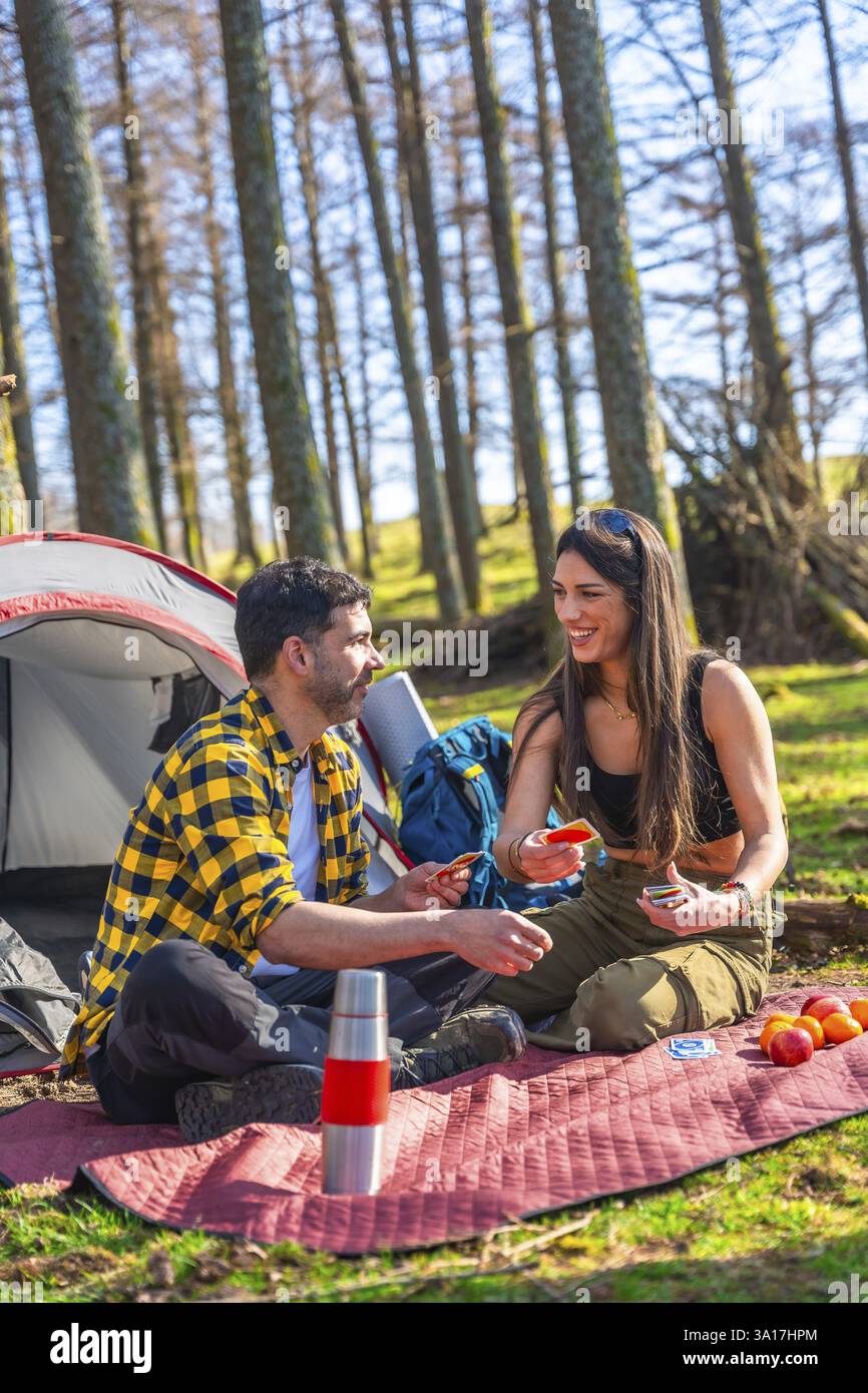Coppia che si gode un rilassante viaggio in campeggio, giocando a carte vicino alla tenda in mezzo alla bellezza serena della foresta e degli alberi Foto Stock
