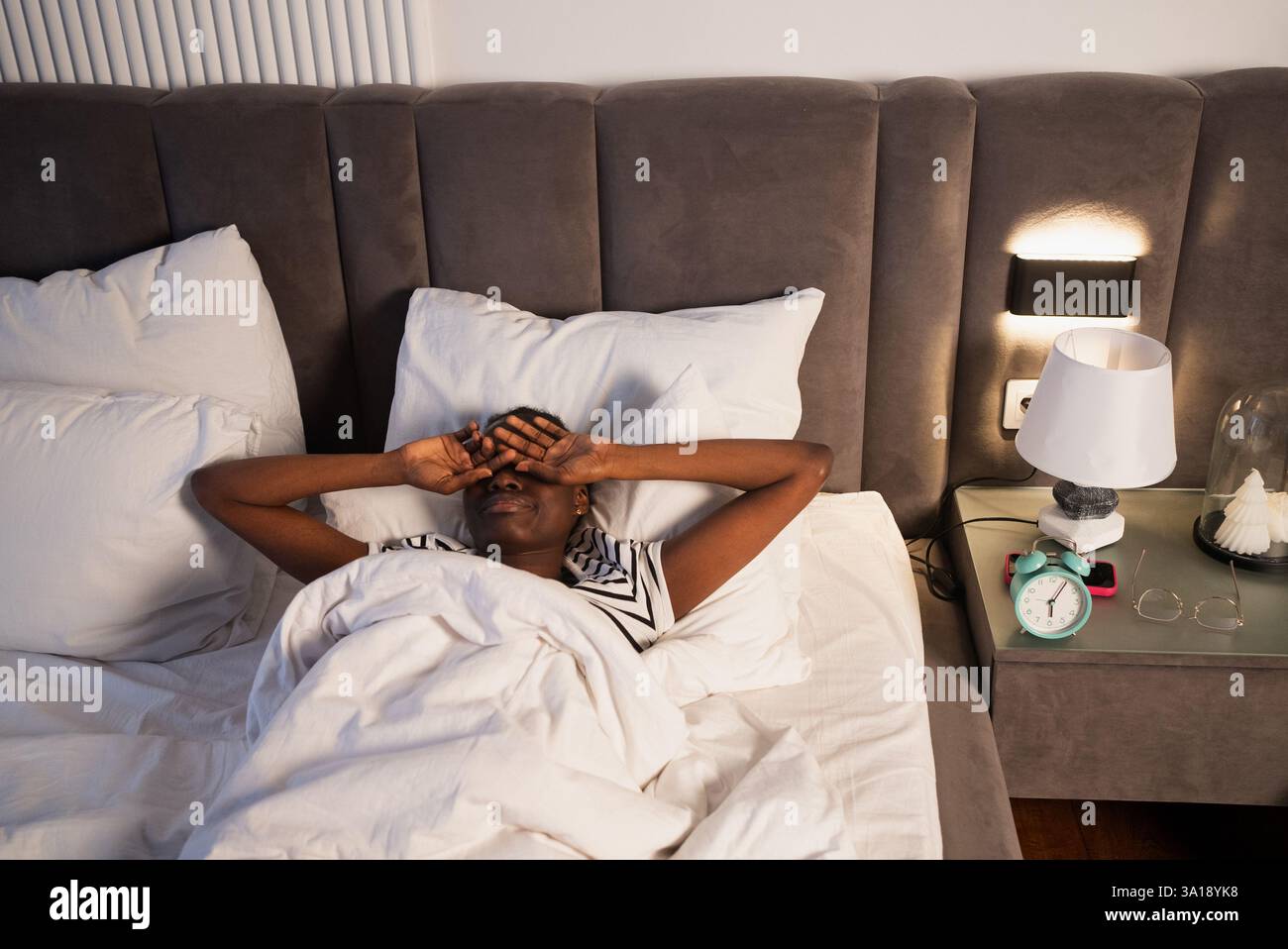 Donna nera che riposa nel letto, che protegge le mani dagli occhi, accoccolata tra morbidi cuscini, comodino con sveglia e lampada, che raffigura un tranquillo relax mattutino o notturno Foto Stock