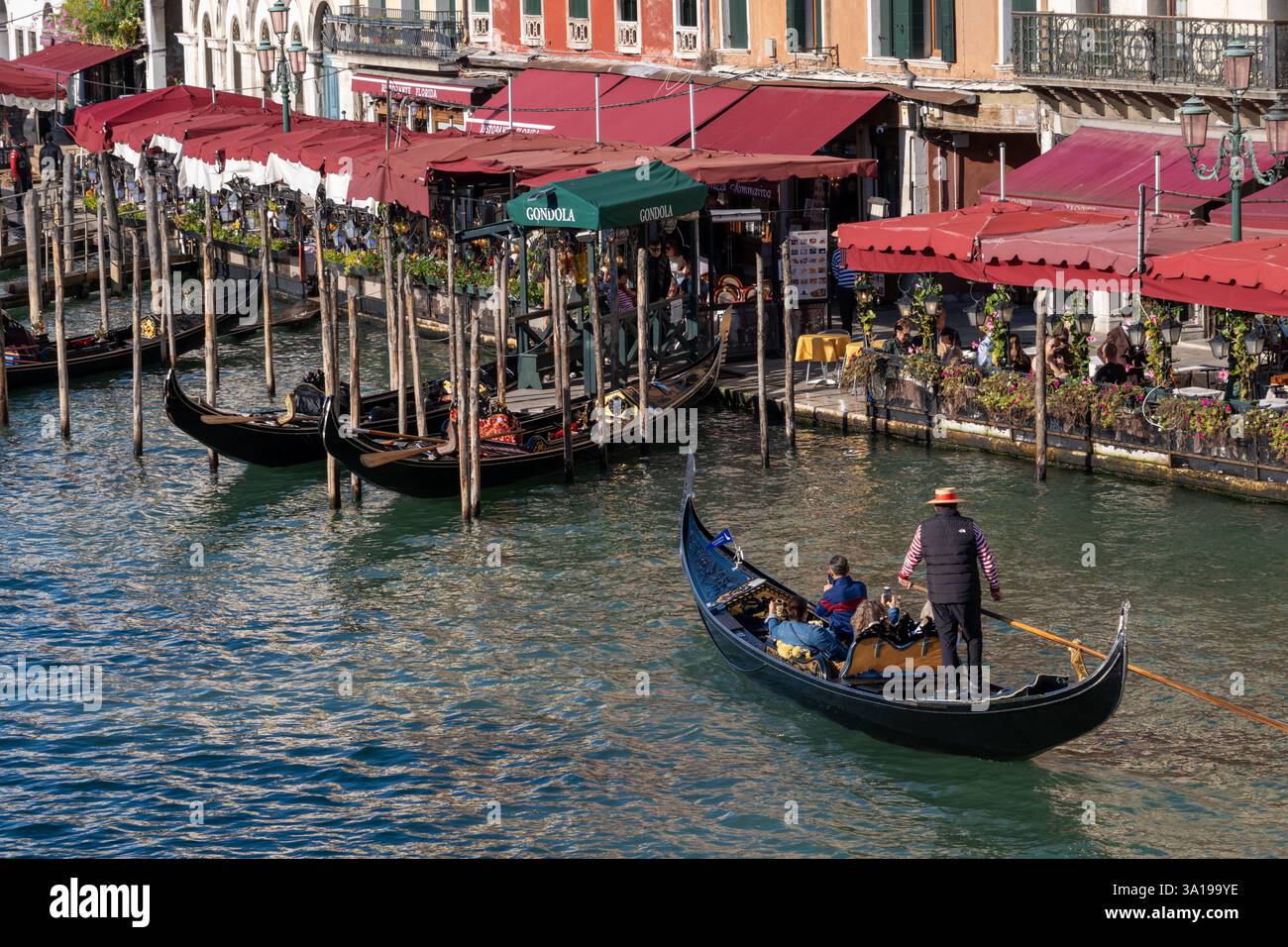 Venezia, Italia - 12 ottobre 2024: Una vivace scena sul lungomare lungo il Canal grande, con le gondole ormeggiate vicino a una fila di affascinanti caffè e ristoranti. Foto Stock
