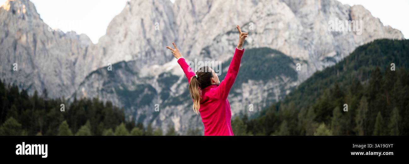 Una donna con una giacca rosa solleva le braccia con gioia sullo sfondo di montagne maestose e foreste lussureggianti, abbracciando la bellezza dell'aria aperta. Foto Stock