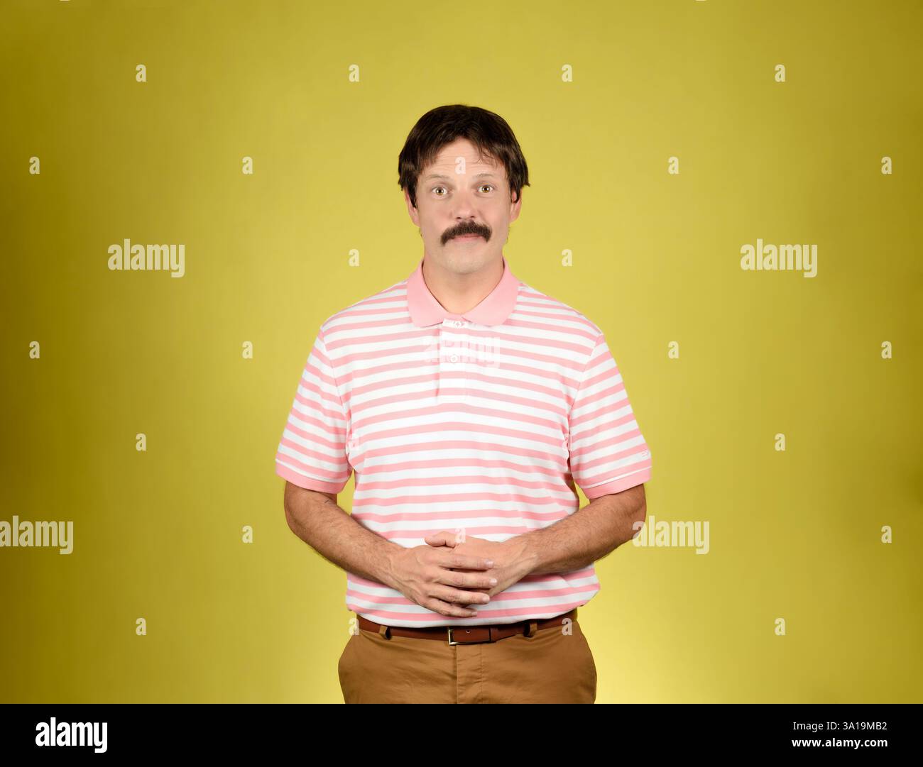 Giovane adulto dai capelli scuri con baffi, polo a righe rosa e bianca, pantaloni marroni, che guardano la macchina fotografica con un sorriso e mani agganciate in franco Foto Stock