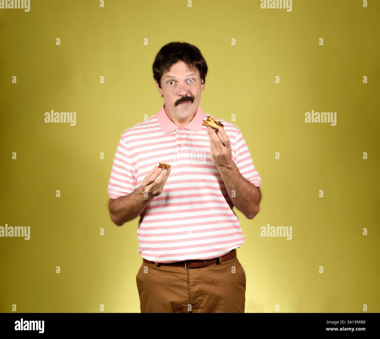 Giovane adulto dai capelli scuri con baffi, polo a righe rosa e bianca, pantaloni marroni, guardare la macchina fotografica e mangiare una torta con una fa soddisfatta Foto Stock