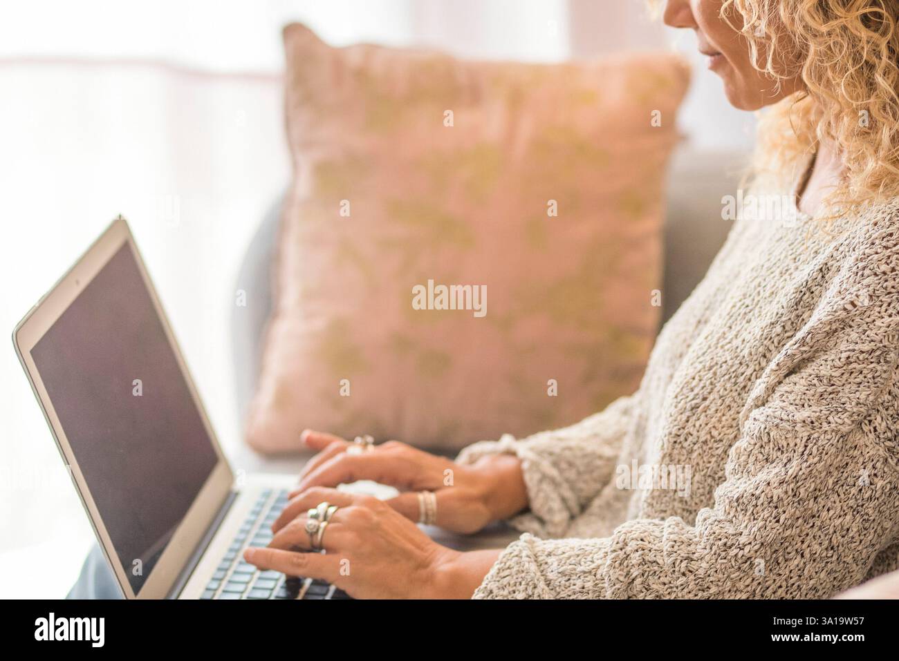 Donna bionda felice con capelli ricci che utilizza un computer portatile seduto sul divano di casa. Donna di mezza età sorridente di 45 anni che guarda il computer mentre naviga su Internet, fa shopping sul sito web e si rilassa sul divano del soggiorno. Foto Stock