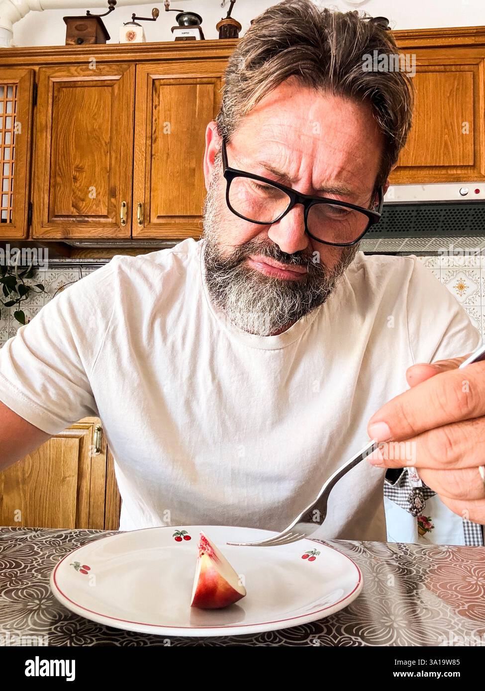 Un adulto arrabbiato con un pranzo dietetico per perdere peso nel ritratto verticale della fotocamera del telefono e in casa in cucina sullo sfondo. Maschio maturo che mangia un pezzo di frutta per cena per fare colazione dietetica senza calorie Foto Stock