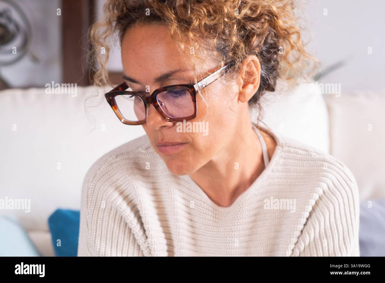 Una donna adulta triste e sconvolta seduta sul divano con le mani strette che guardano in basso. Il concetto di ansia e stress crea problemi alle persone. Una donna a casa sul divano guarda in basso. Speranza e tristezza. Attività ricreative al coperto Foto Stock