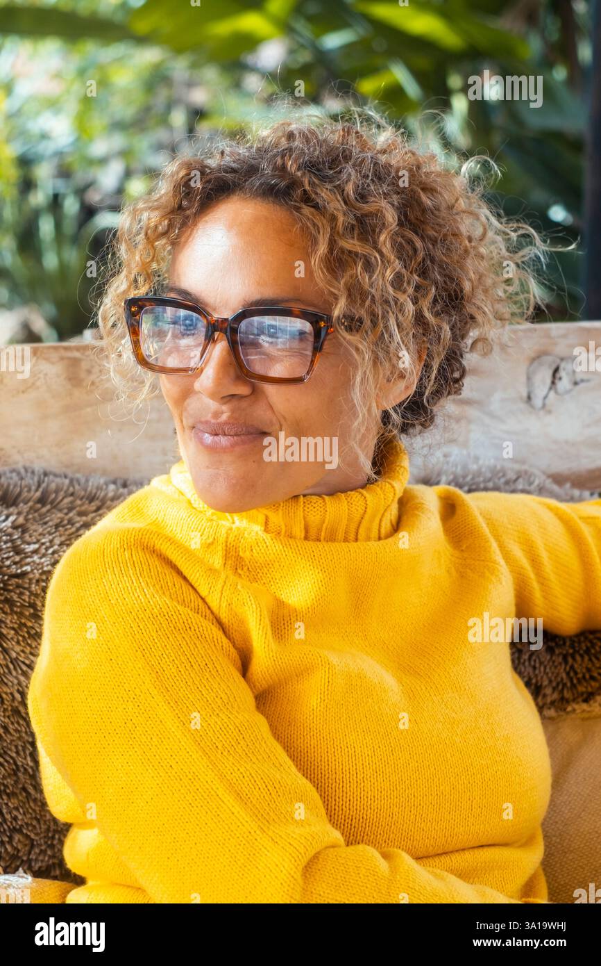 Ritratto di una donna allegra e serena che si rilassa nel giardino seduto e sorridente. Donna che indossa occhiali e pullover giallo. Persone reali all'aperto attività di vita da sole. Donna sicura di sé Foto Stock