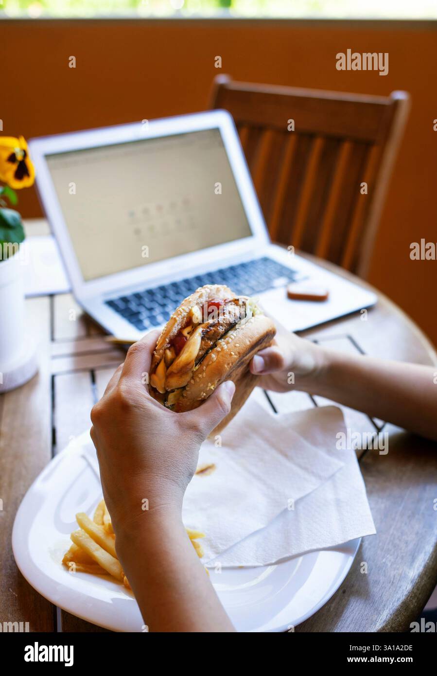 Primo piano delle mani di una donna, mentre mangia un cheeseburger durante la pausa pranzo, nel periodo di quarantena a causa del coronavirus covid-19. Un autentico momento di lavoro intelligente con un notebook a casa. Foto Stock