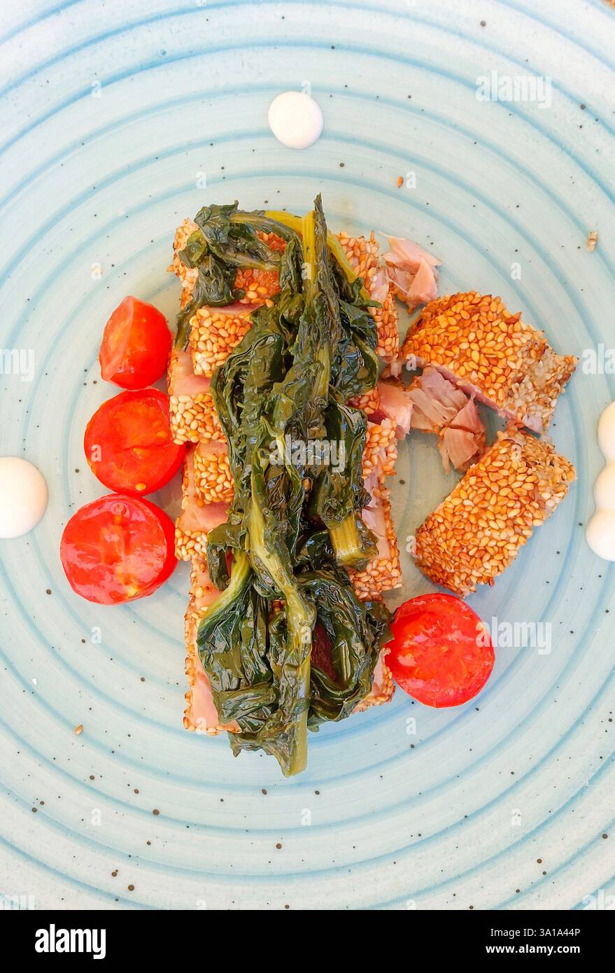 Tonno sfrigolante in crosta di sesamo abbinato a scarola saltata e pomodori ciliegini su un piatto blu vivace Foto Stock