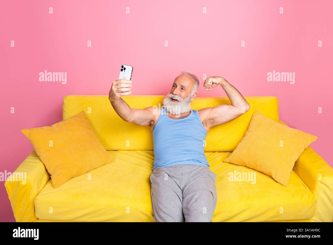 Uomo maturo con braccio flessibile per la barba e sorridente mentre scatta selfie su smartphone da un divano giallo su sfondo rosa in interni Foto Stock