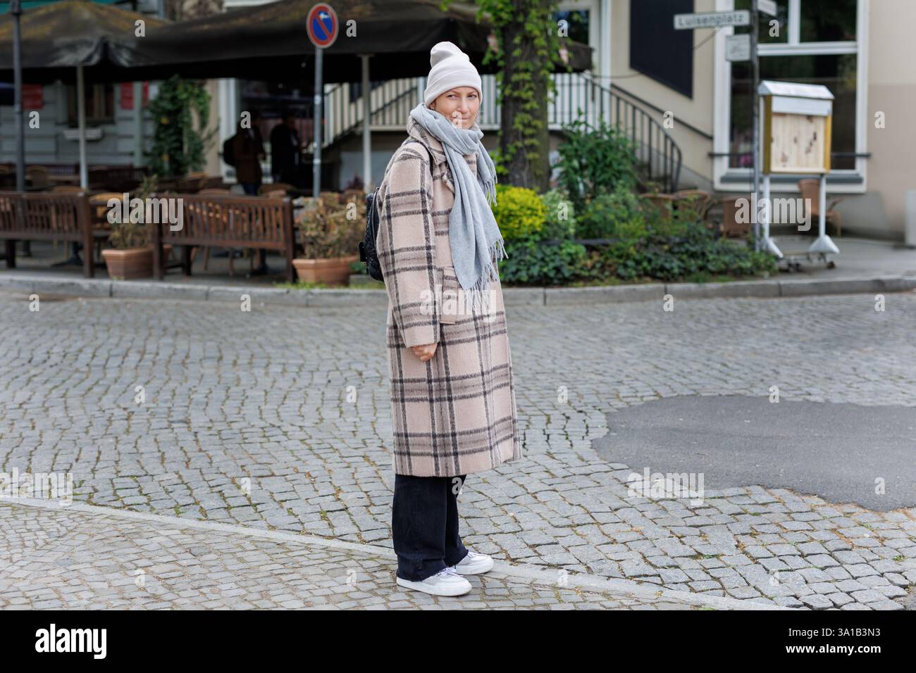 Ritratto di una donna dai vestiti caldi, indossa una sciarpa e un cappello a scacchi in una strada acciottolata a Potsdam. Confuso dubbio incerto divertente facciale Foto Stock