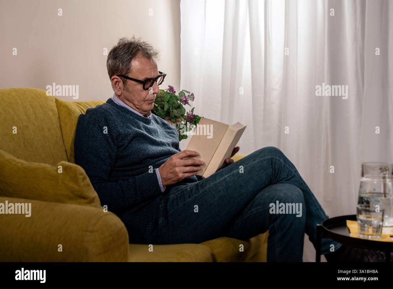 Uomo anziano con occhiali che leggono un libro seduto su un divano giallo in un ambiente accogliente. Un tranquillo momento di relax con una luce calda. Foto Stock